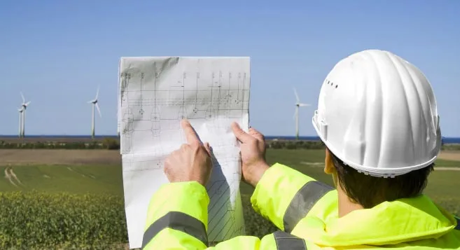 Ingenieur mit PSA und Bauplan schaut auf onshore Windkraftanlagen bei Sonnenschein
